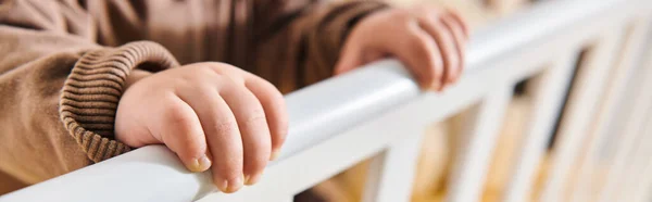 Childhood, tiny hands of little kid standing in crib in nursery room at home, horizontal banner — Stock Photo