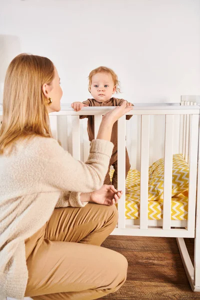 Mãe olhando adorável bebê menino de pé no berço no quarto do berçário em casa, maternidade feliz — Fotografia de Stock