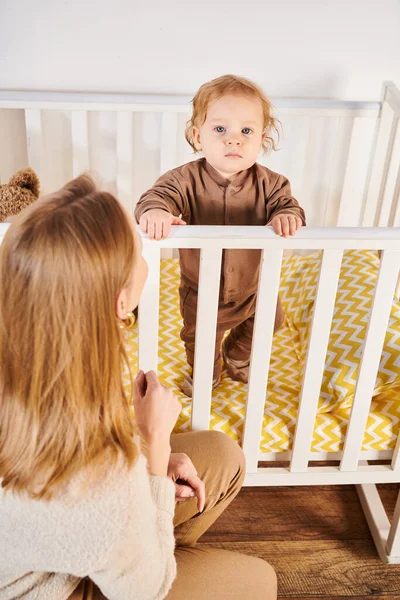 Mulher olhando bonito criança filho de pé no berço no quarto do berçário em casa, maternidade feliz — Fotografia de Stock