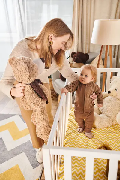 Woman holding teddy bear near cute little son standing in crib in nursery room, happy motherhood — Stock Photo