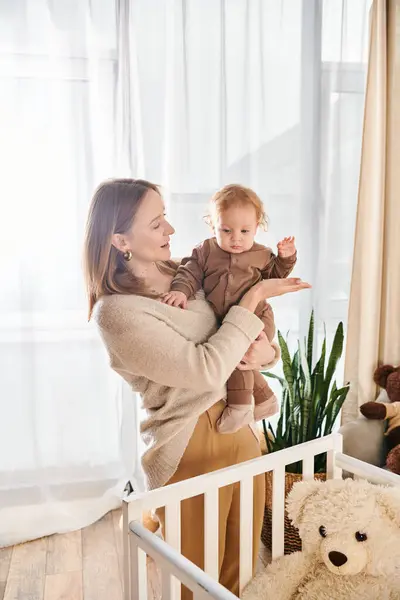 Mujer alegre sosteniendo pequeño hijo en las manos cerca de cuna en cuarto de la guardería en el hogar, maternidad dichosa - foto de stock