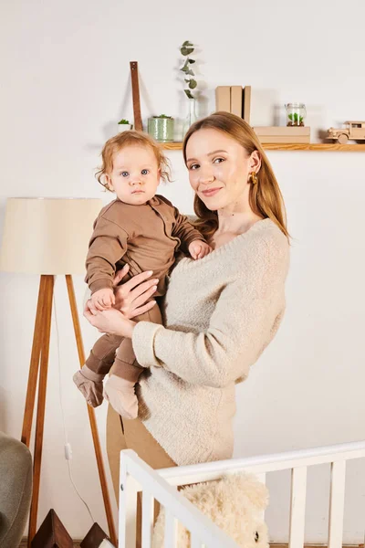 Alegre mujer con adorable toddle hijo en manos mirando a cámara cerca cuna en cuarto de vivero en casa - foto de stock