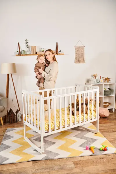 Jovem feliz com o filho pequeno bonito em mãos de pé perto berço no quarto do berçário, maternidade — Fotografia de Stock