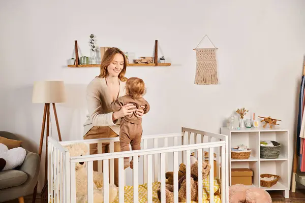 Joven madre alegre sosteniendo pequeño hijo pequeño cerca de cuna en cuarto de la guardería, maternidad dichosa - foto de stock