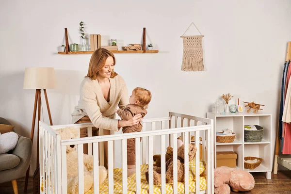 Радостная молодая мать держит милого малыша сына рядом с кроваткой в детской комнате, блаженное материнство — стоковое фото