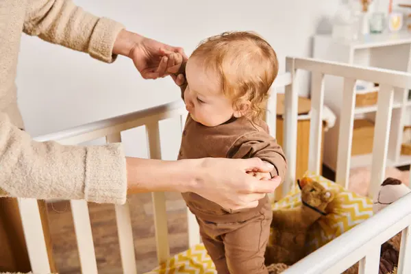 Jeune femme tenant la main d'un petit fils debout dans un berceau dans une chambre d'enfant à la maison, soins et soutien — Photo de stock