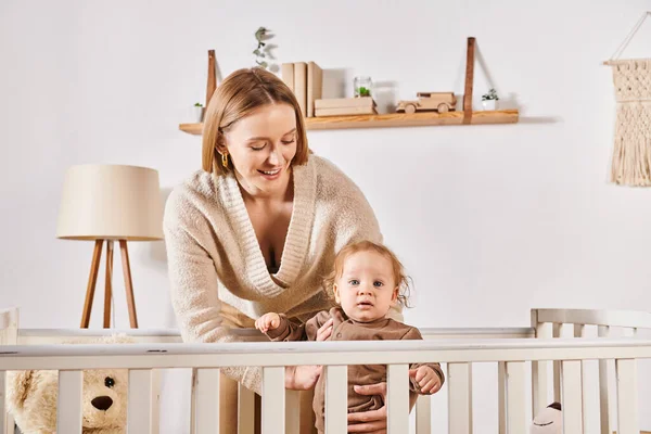 Smiling woman embracing adorable son standing in crib in nursery room at home, care and support — Stock Photo