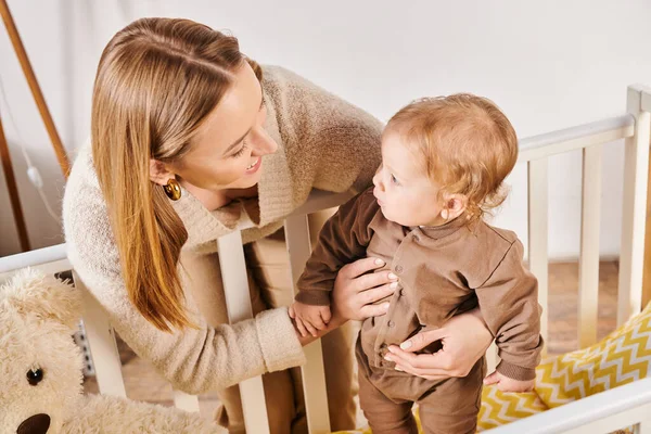 Felice madre sostenere adorabile bambino in piedi in culla nella stanza dei bambini a casa, banner — Foto stock