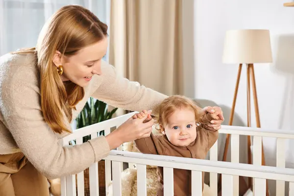 Smiling mother holding hands of cute little son standing in crib in nursery room, love and care — Stock Photo