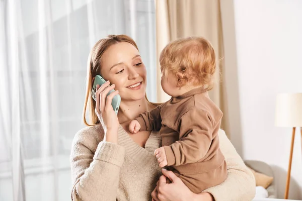 Mujer feliz sosteniendo al hijo en las manos y hablando por teléfono móvil en la habitación de la guardería, madre multitarea - foto de stock