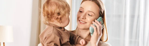 Alegre madre multitarea sosteniendo al hijo en las manos y hablando en el teléfono inteligente en la habitación de la guardería, pancarta - foto de stock