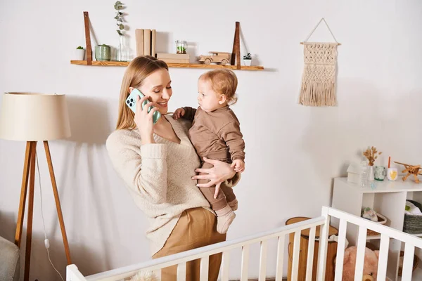 Joyful woman with toddler kid in hands talking on mobile phone near crib, multitasking mother — Stock Photo