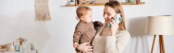 Улыбающаяся женщина с малышом-сыном в руках разговаривает по мобильному телефону в детской комнате, горизонтальный баннер — стоковое фото
