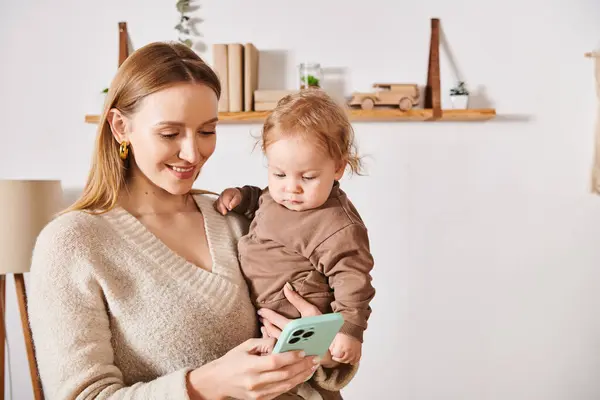 Mãe alegre com menino nas mãos mensagens no telefone celular no quarto de crianças, mulher multitarefa — Fotografia de Stock