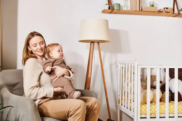 Young woman with adorable son sitting in armchair near crib in nursery room, happy motherhood — Stock Photo