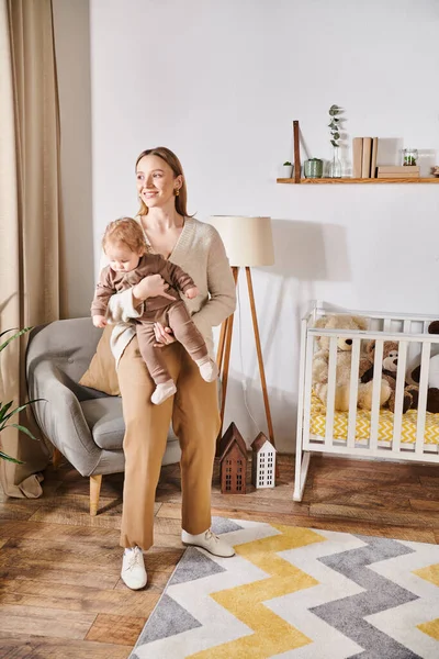 Mãe alegre segurando o filho pequeno adorável perto do berço na poltrona no quarto de quarto de crianças, parentalidade moderna — Fotografia de Stock