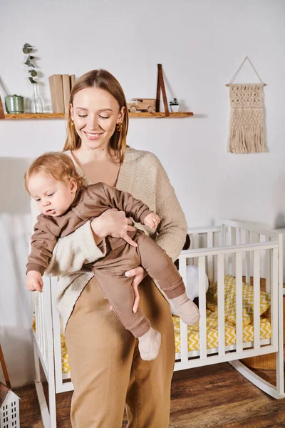 Joyful mother holding cute little son near crib in cozy nursery room, happy modern parenting — Stock Photo