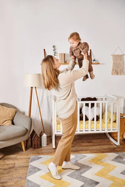 Young woman holding little son while playing and having fun near crib in nursery room, motherhood — Stock Photo