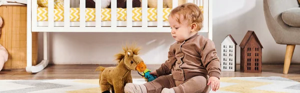 Bonito menino brincando com brinquedo cavalo perto berço no chão no quarto de berçário, bandeira horizontal — Fotografia de Stock
