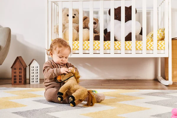 Bonito criança menino sentado no chão e brincando com brinquedo cavalo perto berço no quarto acolhedor berçário — Fotografia de Stock