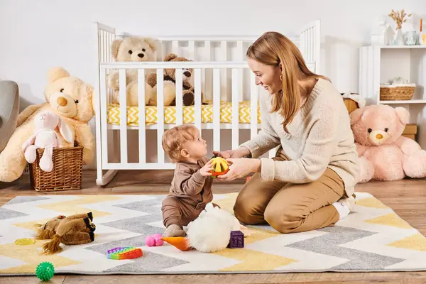 Madre feliz y niño pequeño jugando con juguetes blandos cerca de la cuna en la habitación de la guardería, crianza moderna - foto de stock