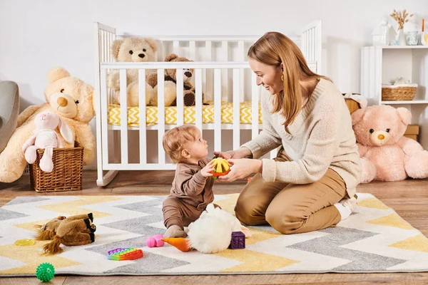 Happy mother and toddler child playing with soft toys near crib in nursery room, modern parenting — Stock Photo