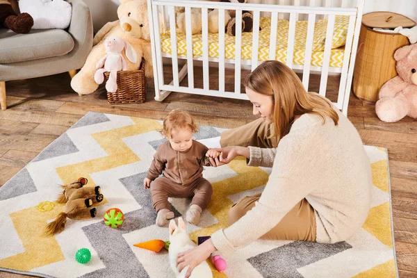 Радостная мать и ребенок сын играет с мягкими игрушками возле кроватки в детской комнате, современное воспитание — стоковое фото