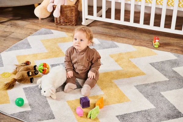 Criança adorável sentado no chão perto de brinquedos macios e berço no quarto de berçário acolhedor, infância feliz — Fotografia de Stock