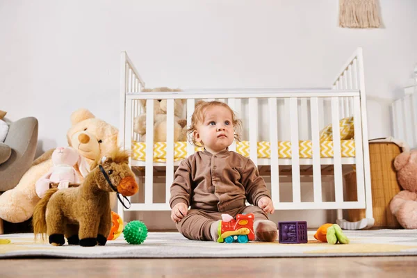 Menino inocente sentado no chão perto berço e brinquedos macios em quarto de berçário acolhedor, bem-aventurado babyhood — Fotografia de Stock