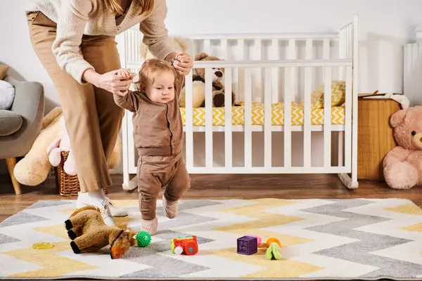 Madre joven cogida de la mano del pequeño hijo caminando cerca de juguetes y cuna en la habitación del vivero, apoyo - foto de stock