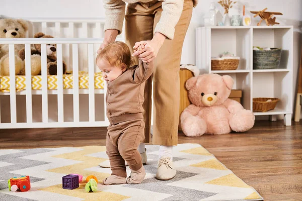 Madre cogida de la mano del hijo pequeño caminando cerca de juguetes y cuna en la habitación del vivero, cuidado y apoyo - foto de stock