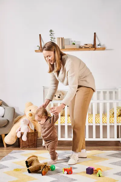 Smiling mother holding hands of baby boy walking near toys and crib in nursery room, support — Stock Photo