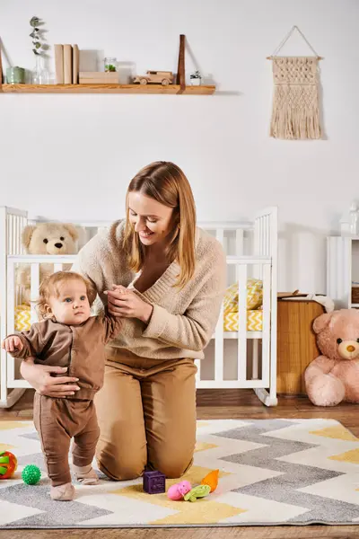 Alegre madre apoyando a su hijo pequeño caminando cerca de juguetes y cuna en la habitación del vivero, maternidad - foto de stock