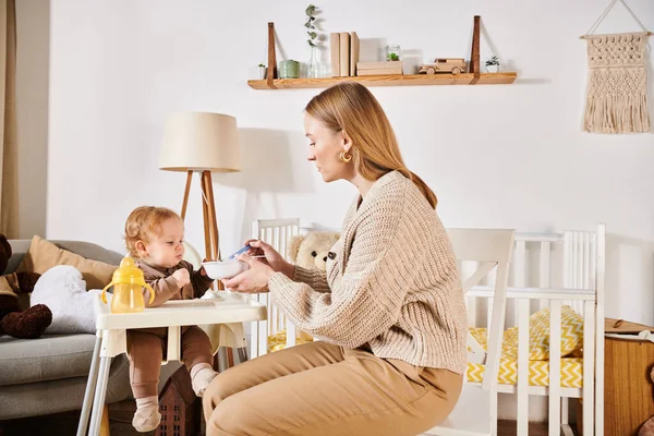 Young mother feeding little son with breakfast on baby chair in nursery room, modern parenting — Stock Photo