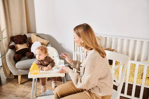 Young woman feeding toddler son with breakfast on baby chair in nursery room, modern parenting — Stock Photo