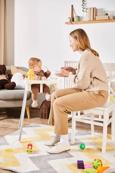 Mulher feliz alimentando filho criança com café da manhã na cadeira do bebê no quarto do berçário, maternidade feliz — Fotografia de Stock