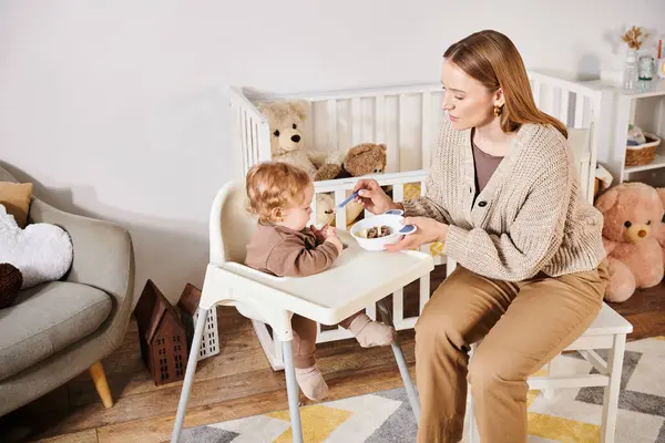 Caring woman feeding baby boy with breakfast on baby chair in nursery room, blissful motherhood — Stock Photo