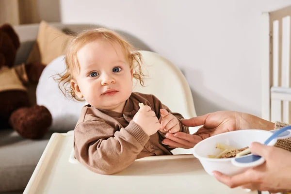 Восхитительный мальчик, сидящий в детском кресле и смотрящий на камеру рядом с мамой с миской завтрака — стоковое фото