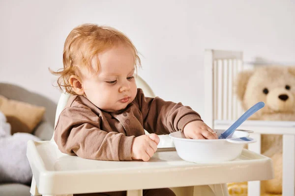 Adorable toddler boy sitting in baby chair near bowl with breakfast in nursery room, childhood — Stock Photo