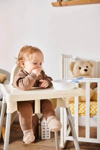 Mignon petit enfant assis dans la chaise de bébé près du bol avec petit déjeuner dans la chambre maternelle, enfance heureuse — Photo de stock