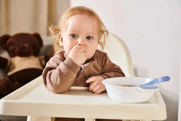 Menino bonito da criança na cadeira do bebê olhando para a câmera perto da tigela com café da manhã no quarto acolhedor do berçário — Fotografia de Stock