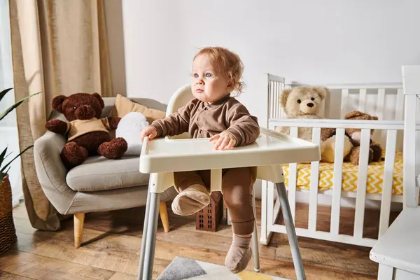 Menino criança sentado em cadeira de bebê e olhando para longe no quarto de berçário acolhedor com berço e ursinhos de pelúcia — Fotografia de Stock
