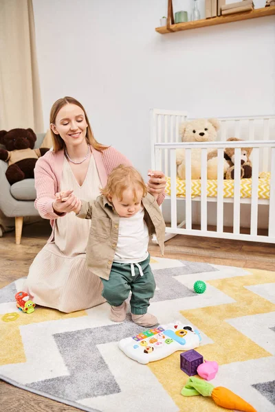 Mujer alegre cogida de la mano del pequeño hijo aprendiendo a caminar en el suelo en la sala de estar en casa, apoyo - foto de stock