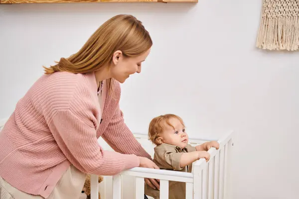 Улыбающаяся женщина, поддерживающая сына малыша, стоящего в кроватке в детской комнате дома, счастливое материнство — стоковое фото