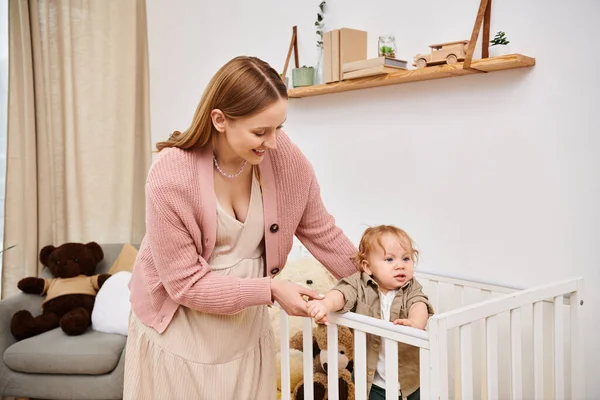 Веселая женщина поддерживает малыша сына стоя в кроватке в детской комнате дома, блаженное материнство — стоковое фото