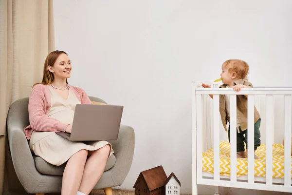 Smiling woman working on laptop near toddler son in baby crib in nursery room, multitasking mother — Stock Photo
