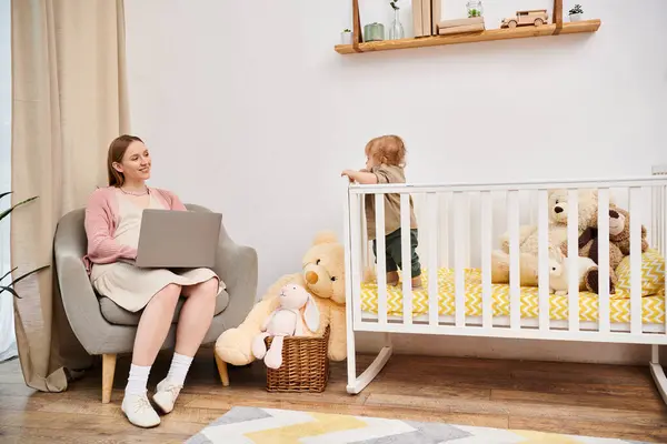 Радостная женщина работает на ноутбуке рядом с маленьким сыном в детской кроватке в детской комнате, многозадачная мать — стоковое фото