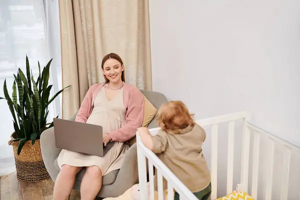 Sonriente mujer trabajando en el ordenador portátil cerca de niño pequeño hijo en cuna en la habitación de la guardería, multitarea - foto de stock
