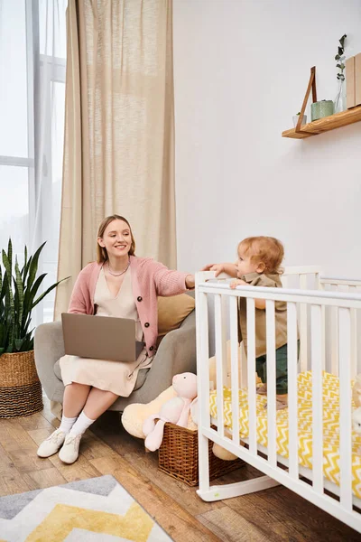 Mujer alegre que trabaja en el ordenador portátil cerca de un niño lindo en la cuna en la habitación de la guardería, madre multitarea - foto de stock