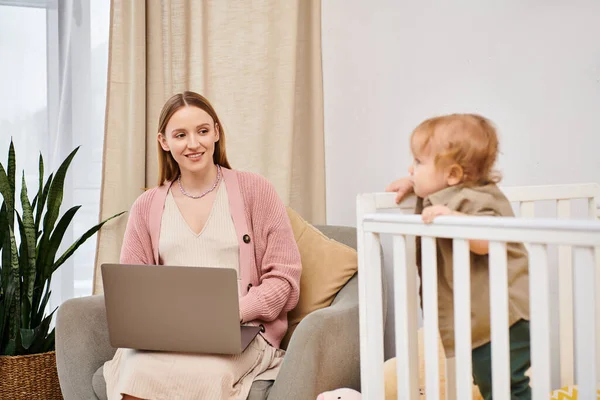 Mère heureuse travaillant sur ordinateur portable près de mignon enfant dans la crèche de bébé dans la chambre maternelle, femme multitâche — Photo de stock